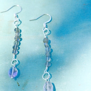 Puede incluir: Un par de pendientes colgantes de plata con un diseño de piedras preciosas gris claro y púrpura.