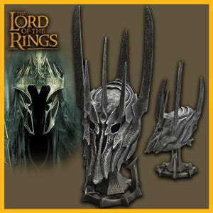 May include: A silver-colored replica of the One Ring from The Lord of the Rings, with a detailed design of spikes and a skull-like face. The ring is displayed on a black stand with a circular base.