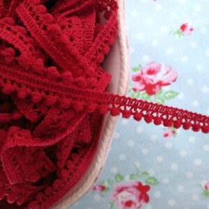 May include: Red pom-pom trim with a crocheted design. The trim is in a bowl and a piece is hanging over the edge.