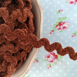 May include: A close-up of brown, fuzzy, wavy trim ribbon. The ribbon is in a white bowl and on a blue and white floral fabric background.