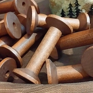 May include: A collection of multiple empty wooden spools with a medium brown finish. Each spool features a cylindrical central shaft and two circular end flanges with a small hole in the center, arranged in a scattered pile with various orientations.
