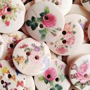 May include: Assortment of round wooden buttons with floral designs. The buttons are white with pink, yellow, and green floral patterns.