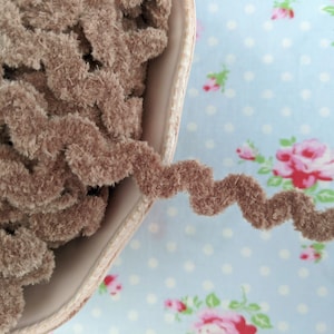 May include: A close-up of a brown, fuzzy, curly trim. The trim is in a white bowl and is spilling out onto a blue and white floral fabric.