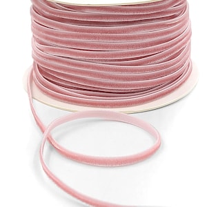 May include: A spool of dusty rose velvet ribbon. The ribbon is tightly wound on a cardboard core, with a length of ribbon unwound and lying in front. The ribbon has a soft, plush texture and a delicate, muted pink color.