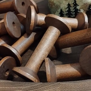 May include: A collection of multiple empty brown wooden spools for thread, each featuring a cylindrical center and two circular end flanges with a central hole. The spools are arranged in a scattered pile, showing the natural wood grain texture.