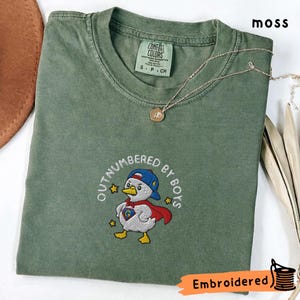 May include: A moss green t-shirt featuring an embroidered cartoon duck in a superhero outfit. The text "OUTNUMBERED BY BOYS" is embroidered around the duck. A gold necklace with a pendant is placed on the shirt.