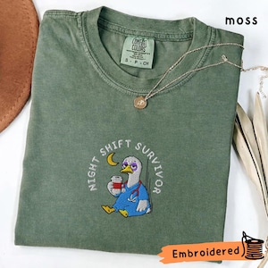 Peut inclure: T-shirt vert mousse avec la phrase brodée "Night Shift Survivor" autour d'un canard de dessin animé portant une blouse et tenant une tasse de café. Un collier doré et le mot "Embroidered" sont également visibles.