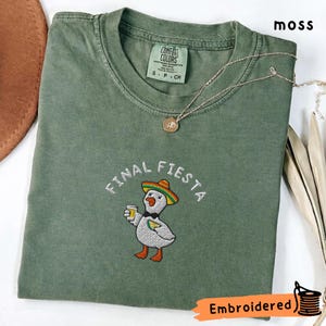 May include: A moss green t-shirt with the words "FINAL FIESTA" embroidered above a cartoon duck wearing a sombrero and holding a drink. A gold necklace with a pendant is draped across the shirt. The shirt has a Comfort Colors tag.
