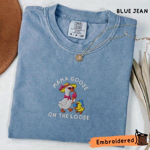 May include: A blue jean coloured t-shirt featuring an embroidered graphic of a goose wearing a yellow hat and pink sunglasses holding an ice cream cone, with a small duckling beside it. The text reads MAMA GOOSE ON THE LOOSE in white thread.