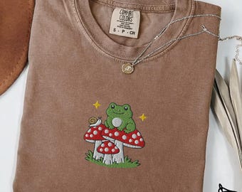 May include: Espresso-colored t-shirt with an embroidered design of a green frog and a snail on a red and white mushroom. The shirt has a Comfort Colors label and a silver necklace. The word "Embroidered" is at the bottom.