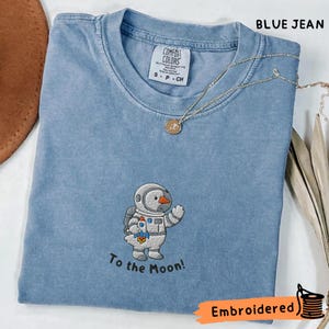 Puede incluir: Camiseta azul vaquero con un diseño bordado de un muñeco de nieve astronauta y el texto "To the Moon!". La camiseta tiene cuello redondo y una pequeña etiqueta en el cuello. Un collar dorado con un colgante está sobre la camiseta.