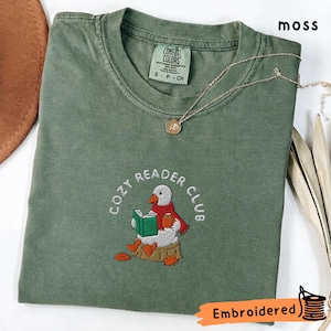 May include: A moss green t-shirt with an embroidered design of a duck reading a book, wearing a scarf, and holding a mug. The text "COZY READER CLUB" is arched above the duck. A gold necklace rests on the shirt.