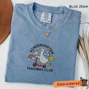 以下が含まれることがあります： 赤いスクーターに乗った白いアヒルの刺繍が施されたブルーデニムカラーのTシャツ。デザインには虹、クレヨン、アルファベットの積み木、黄色い星が含まれており、その下に「KINDERGARTEN TEACHERS CLUB」という文字が刺繍されています。