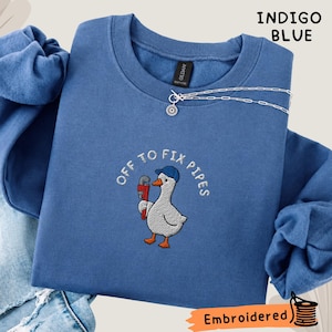 May include: An indigo blue crewneck sweatshirt featuring an embroidered graphic of a white goose wearing a blue cap and holding a red pipe wrench. The text "OFF TO FIX PIPES" is arched above the goose. A silver chain necklace rests on the collar.