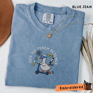 以下が含まれることがあります： 青いジーンズ色のTシャツで、青いゴーグルをつけた白いガチョウが青いスクーターに乗っている刺繍が施されています。テキストは「OFF TO TEACH SCIENCE」と書かれており、原子、試験管、ペトリ皿、顕微鏡が周囲に配置されています。