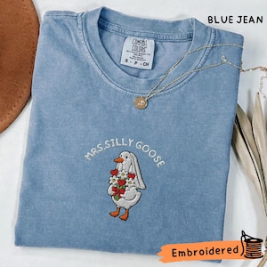 Puede incluir: Camiseta azul vaquero con la frase bordada "Mrs. Silly Goose" y un ganso sosteniendo flores. La camiseta tiene cuello redondo y mangas cortas. Un collar dorado con un colgante está drapeado sobre la camiseta.