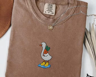 May include: Espresso-colored t-shirt with an embroidered design of a white goose wearing yellow boots and carrying carrots. The shirt has a crew neck and short sleeves. A silver necklace with a pendant is draped across the shirt. The word "Embroidered" is at the bottom.