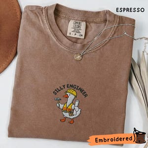 以下が含まれることがあります： 「SILLY ENGINEER」の刺繍と、ヘルメットをかぶり、ハンマーと設計図を持った漫画のガチョウが描かれたエスプレッソカラーのTシャツ。 シャツにはComfort Colorsのタグと小さなシルバーのネックレスが付いています。