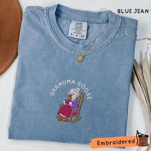 Puede incluir: Camiseta azul vaquero Comfort Colors con un diseño bordado de un ganso en una mecedora, sosteniendo una taza. El texto "Grandma Goose" está arqueado sobre el ganso. Un collar dorado con un colgante está en la camisa.