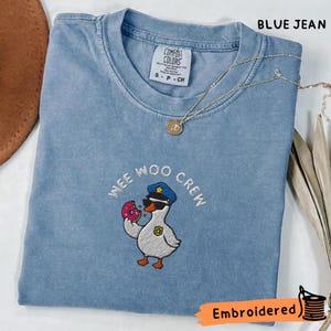 Op de afbeelding: Een blauw spijkerkleurig t-shirt met een geborduurde afbeelding van een eend met een politiepet en badge, die een roze donut vasthoudt. De tekst WEE WOO CREW is in een boog boven de eend geborduurd. Een dunne gouden ketting ligt op het t-shirt.