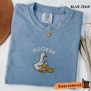 May include: Blue jean Comfort Colors t-shirt with the word "HOOKER" embroidered above a cartoon goose knitting. The goose is knitting with yellow yarn. A gold necklace with a pendant is draped on the shirt. The word "Embroidered" is in the bottom right corner.