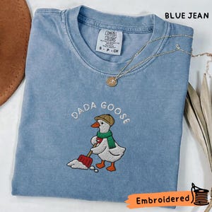 Peut inclure: Un t-shirt bleu jean à col rond avec une broderie représentant une oie portant un chapeau beige et une écharpe verte, tenant une pelle rouge. Le texte DADA GOOSE est brodé au-dessus de l'oie. Un fin collier à chaîne dorée repose sur le t-shirt.