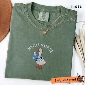 Peut inclure: Un t-shirt à manches courtes vert mousse avec une broderie représentant un canard blanc portant un stéthoscope bleu et tenant un caneton enveloppé dans une couverture bleue. Le texte NICU NURSE est brodé en arc au-dessus du canard.