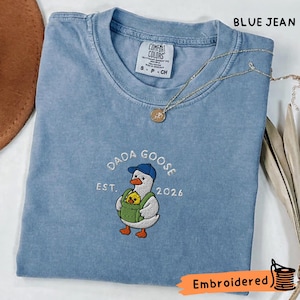 以下が含まれることがあります： 青いジーンズ色のTシャツで、青い帽子をかぶり、黄色いひよこが入った緑色の抱っこ紐をつけた白いガチョウの刺繍が施されています。ガチョウのイラストの上と横には「DADA GOOSE EST. 2026」という文字が刺繍されています。