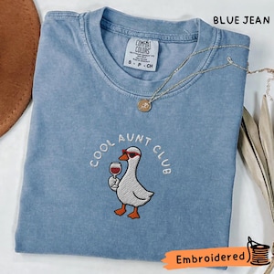 Op de afbeelding: Een blauw spijkerkleurig t-shirt met een geborduurde afbeelding van een witte gans met een rode zonnebril die een glas rode wijn vasthoudt. De tekst COOL AUNT CLUB staat in een boog boven de gans. Het shirt heeft een ronde hals en een Comfort Colors label.