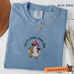 May include: A blue jean coloured t-shirt featuring an embroidered graphic of a goose wearing a pink unicorn headband and sunglasses, holding a teddy bear and a coffee cup. The text reads IN MY GIRL DAD ERA. The shirt has a Comfort Colors brand label.