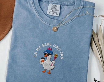 May include: A blue jean Comfort Colors t-shirt with an embroidered design. The design features a cartoon goose wearing a hat and holding a coffee cup and a magic wand. The text "IN MY GIRL DAD ERA" is embroidered around the goose.