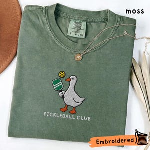 Könnte beinhalten: Ein moosgrünes T-Shirt mit einer Stickerei, die eine weiße Gans mit einem grün-weißen Pickleball-Schläger und einem gelben Ball zeigt. Der Text "PICKLEBALL CLUB" ist unter der Gans eingestickt. Das T-Shirt hat ein Comfort Colors Etikett.