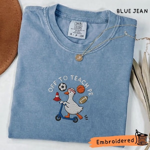 以下が含まれることがあります： 青いジーンズ色のTシャツで、赤いヘッドバンドをして青いスクーターに乗った白いアヒルの刺繍が施されています。OFF TO TEACH PEという文字の周りには、サッカーボール、バスケットボール、フットボール、赤い三角コーンが描かれています。