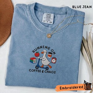以下が含まれることがあります： 青いジーンズ色のTシャツで、眼鏡をかけたガチョウが青いスクーターに乗っている刺繍が施されています。デザインには、赤いリンゴ、本の山、コーヒーカップ、目覚まし時計が含まれており、RUNNING ON COFFEE & CHAOSという文字が添えられています。