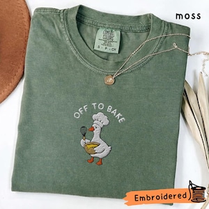 May include: A moss green short-sleeve t-shirt featuring an embroidered graphic of a white goose wearing a chef hat, holding a whisk and a yellow mixing bowl. The text "OFF TO BAKE" is embroidered in white above the goose. The shirt has a crew neck.