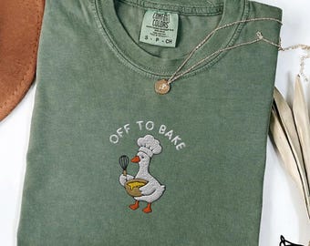 May include: A moss green short-sleeve t-shirt featuring an embroidered graphic of a white goose wearing a chef hat, holding a whisk and a yellow mixing bowl. The text "OFF TO BAKE" is embroidered in white above the goose. The shirt has a crew neck.