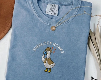 May include: Blue jean colored t-shirt with the embroidered phrase "Sherlock Honks" and a cartoon goose wearing a detective hat and holding a magnifying glass. The shirt has a crew neck and short sleeves. A gold necklace is draped across the shirt.