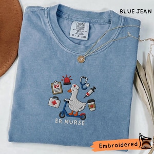 以下が含まれることがあります： ナースキャップを被り青いスクーターに乗った白いガチョウの刺繍が施された、ブルーデニム色のTシャツ。ガチョウの周りには、ERクリップボード、サイレン、聴診器、注射器、コーヒーカップなどの医療用アイコンが刺繍されています。