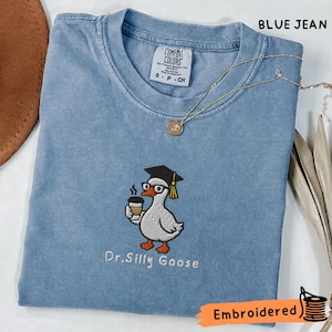 Könnte beinhalten: Ein blaues T-Shirt in Jeansoptik mit einer Stickerei einer weißen Gans, die eine schwarze Doktorhut-Mütze und eine Brille trägt und eine Kaffeetasse hält. Der Text Dr. Silly Goose ist unter der Gans gestickt. Das Shirt hat ein Comfort Colors Etikett.