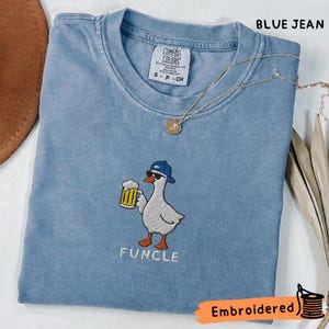 Op de afbeelding: Een blauw spijkerkleurig t-shirt met een geborduurde afbeelding van een witte gans met een blauwe honkbalpet en zonnebril die een bierpul vasthoudt. De tekst "FUNCLE" is onder de gans geborduurd. Het shirt heeft een Comfort Colors label.