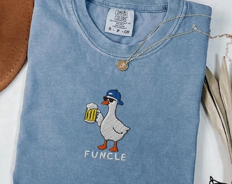 May include: A blue jean coloured t-shirt featuring an embroidered graphic of a white goose wearing a blue baseball cap and sunglasses while holding a beer mug. The text "FUNCLE" is embroidered below the goose. The shirt has a Comfort Colors label.
