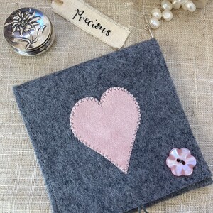 May include: A grey felt pouch with a pink heart applique and a flower-shaped button. A small silver round box with a flower design and a tag that says "Precious" are also in the image. The pouch is closed with a loop.