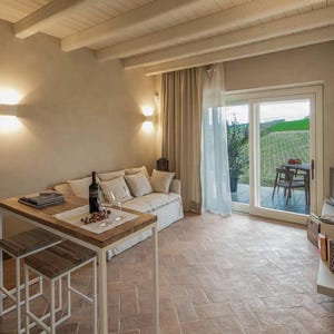Può includere: Un accogliente spazio abitativo con un tavolo e sgabelli in legno, un divano e una porta scorrevole che conduce a un patio. Una bottiglia di vino e bicchieri sono sul tavolo. La stanza ha un'illuminazione calda e una vista su un paesaggio verde.