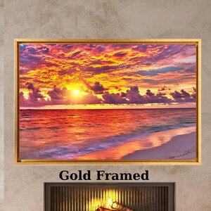 Op de afbeelding: Een rechthoekig wandkunstwerk met een levendige zonsondergang boven de oceaan met oranje, gele en paarse wolken, ingelijst in een dunne gouden lijst. Het kunstwerk hangt boven een open haard met een brandend vuur en een stapel houtblokken.