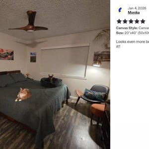 a bedroom with a large bed, a cat resting on the bed, and a painting on the wall. The room has a wooden ceiling fan and a window with a white curtain.