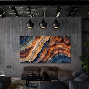 Op de afbeelding: Een groot rechthoekig wandkunstwerk met een gedetailleerd abstract patroon van gelaagde, golvende texturen in tinten diepblauw, gebrand oranje en crème. Het kunstwerk hangt aan een donkergrijze betonnen muur boven een lage bank.