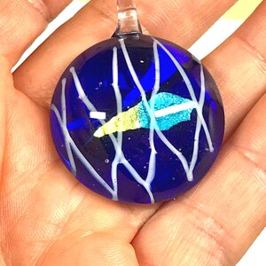 May include: A circular cobalt blue glass pendant featuring a clear glass bail at the top. The surface is decorated with a series of thin, intersecting white glass lines and a central, elongated iridescent accent piece that shifts between yellow and teal.
