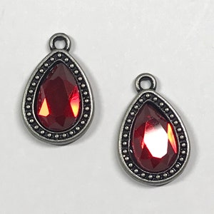 May include: Two teardrop-shaped charms featuring a faceted deep red stone set in an antiqued silver-toned metal frame. Each charm has a small circular loop at the top for attachment, and the metal border is decorated with a beaded texture.