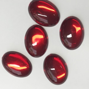 May include: Five smooth, oval-shaped cabochons in a deep, glossy red color. Each piece features a polished, reflective surface with bright white light highlights, showcasing a uniform, rounded dome shape and a consistent, vibrant crimson hue.