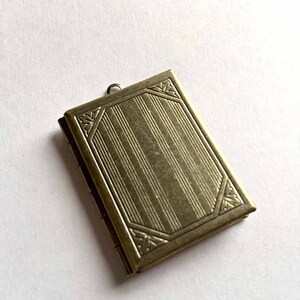 May include: A rectangular antique brass locket featuring a vertical striped pattern on the front cover, framed by decorative corner accents with leaf motifs and a small metal loop at the top for attachment to a chain or cord.